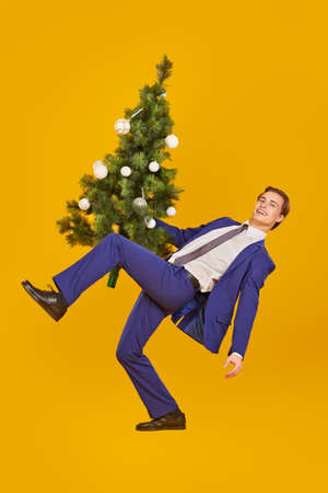 Handsome cheerful young man in elegant formal suit is jumping with a Christmas tree. Yellow background with copy space. Christmas and New Year celebration.の写真素材