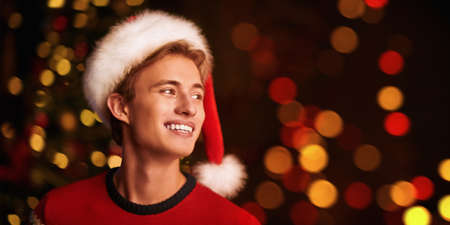 Portrait of a happy handsome young man in Santa Claus hat celebrating Christmas. The backdrop of sparkling holiday lights. Copy space.の写真素材