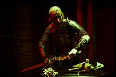 An experienced medieval warrior in armor and with a sword in a wooden house. Stage portrait. The...