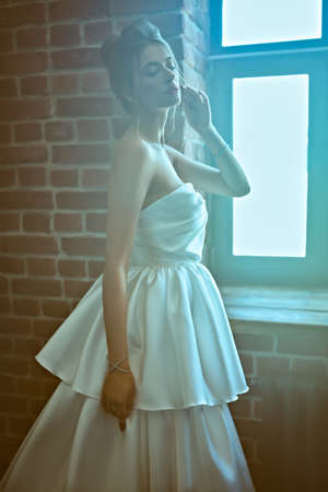 Wedding fashion. Portrait of a beautiful fashion model in a luxury wedding dress standing by the window in a room with loft interior.の写真素材