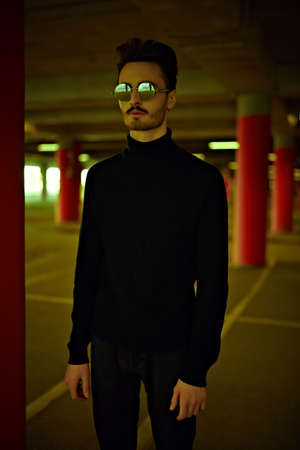 Young man hipster wearing black pullover and round sunglasses stands in the underground Parking lot. Men's fashion.の写真素材