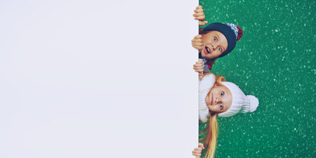 Two beautiful kids in winter clothes look out from behind the white board and are smiling. Green...