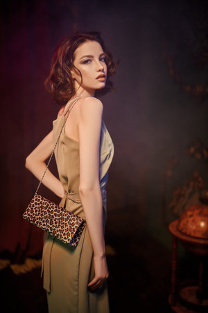 Young elegant woman in evening dress stands in a darkened room with a classic vintage interior.