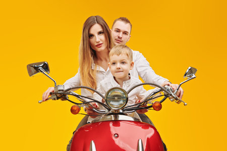Joyful parents and their cute little son ride a red scooter on a bright yellow background. Christmas and New Year celebration.の写真素材