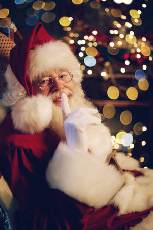 Portrait of a happy Santa Claus showing a secret sign. Beautiful Christmas interior and festive lights are shining around.の写真素材