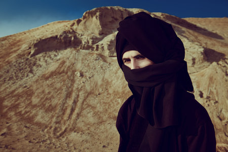 Courageous young man in black clothes and black keffiyeh on his head stands in a desert and looks...