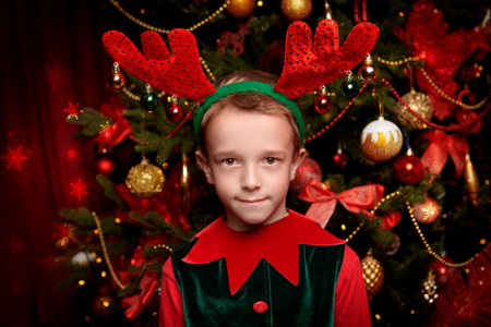 Portrait of a cute elf boy standing by a beautiful Christmas tree.の写真素材