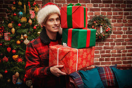 Handsome young man in Santa Claus hat stands with gifts by a beautiful christmas tree and smiles. Christmas and New Year.の写真素材
