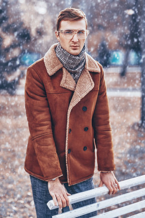 Men's fashion. Handsome young man in winter coat stands by a bench in a city park. Winter, city and people. Clothing and accessories.の写真素材