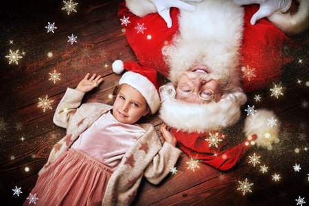 Portrait of a happy little girl and Santa Claus having fun on a floor. Shining lights and snowflakes are around. Merry Christmas and Happy New Year!の写真素材