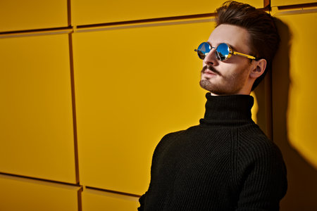 Portrait of a stylish young man in black pullover and round mirror sunglasses posing on a street by the yellow industrial wall. Male fashion. Urban style.の写真素材