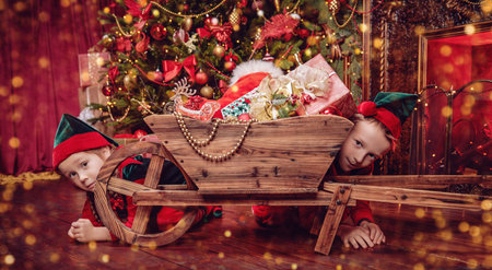 Christmas tale. Little elves are hiding behind a trolley with gifts. They are in a house with a beautiful Christmas interior.の写真素材