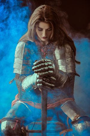 A beautiful noble warrior woman in chain mail and plate armor sits, leaning on her sword and bowing her head. Medieval knight.の写真素材