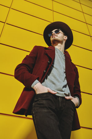 Fashion portrait of an elegant young man in a coat, a hat and sunglasses posing on a street by the yellow industrial wall. Male fashion. Urban style.の写真素材