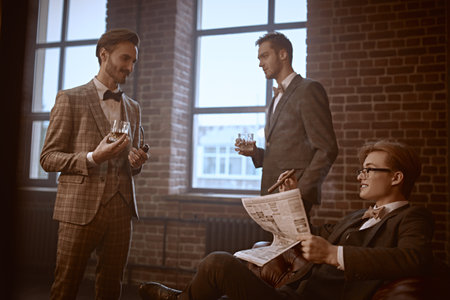 Three handsome men in elegant suits spends time in the men's club with cigars and whiskey. Luxury lifestyle. Men's beauty, fashion.の写真素材