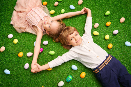 Cute laughing boy and girl are lying on a green lawn with colorful eggs. Easter holiday.