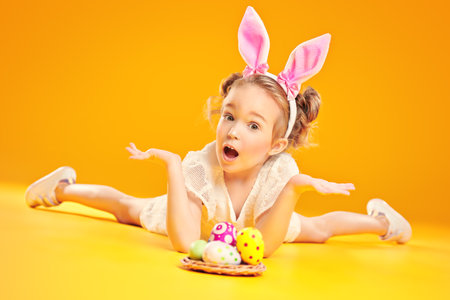Funny emotional child girl with bunny ears poses with painted Easter eggs on a yellow background. Happy childhood. Easter holiday.の写真素材