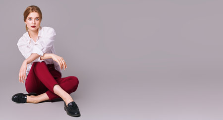Full length portrait of a beautiful blonde woman in white blouse and red pants posing at studio on a gray background. Elegant business style. Copy space.の写真素材