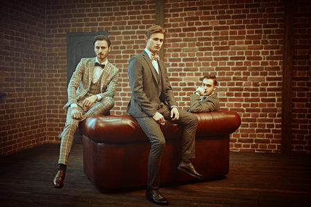 Business people club. Three handsome men in elegant suits sit on a leather sofa in apartments with loft interior. Men's fashion.の写真素材
