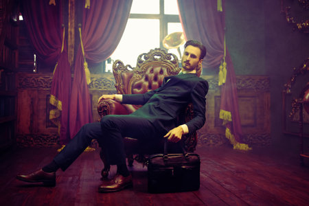 Men's beauty, fashion. Portrait of a handsome man in elegant classic suit sitting in an armchair with a leather bag in a luxury apartment. Luxury lifestyle.の写真素材