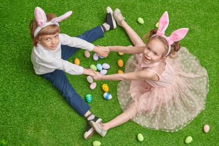 Happy Easter. Joyful boy and a girl with bunny ears are sitting on a green lawn with colorful eggs and smiling.の写真素材