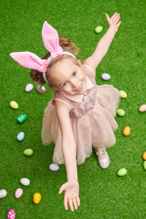 Happy little girl with bunny ears stands on a green lawn with colorful eggs. Easter holiday.の写真素材