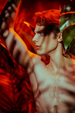 Handsome young man fashion model with naked torso posing among tropical plants in red light. Men's beauty. Perfumery and beauty products.の写真素材