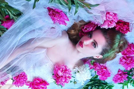 Wedding fashion. Charming brunette woman in a transparent white dress lying among fresh peony flowers. Beauty, make-up and cosmetics.の写真素材