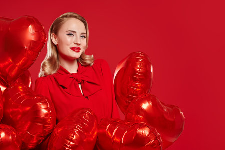 Portrait of a beautiful blonde woman in red dress posing with red heart shaped balloons on a red background. Love and Valentine's Day concept. Beauty, make-up and cosmetics. Copy space.の写真素材