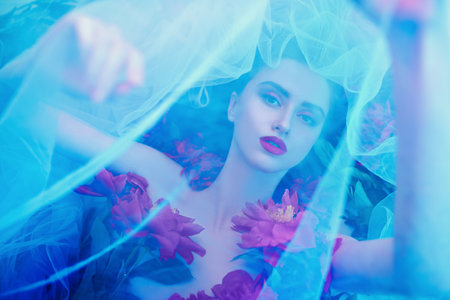 Flowers and beauty. Magnificent brunette woman with bright make-up lying among peony flowers covered with a veil. Cosmetics, make-up. Perfumery.の写真素材