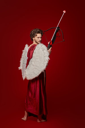 Full length portrait of a handsome Cupid with a crossbow and arrow on a red background. God of Love....