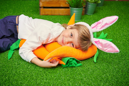 Happy little boy with bunny ears lying on the grass with carrots in a spring garden. Easter holiday.