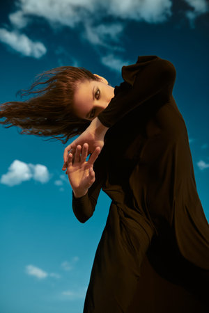 Beautiful expressive fashion model girl in a long black dress poses against a blue sky with white...
