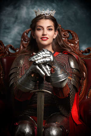 Portrait of a beautiful medieval queen in knightly armor sitting on a throne and holding a sword. History of the Middle Ages.の写真素材