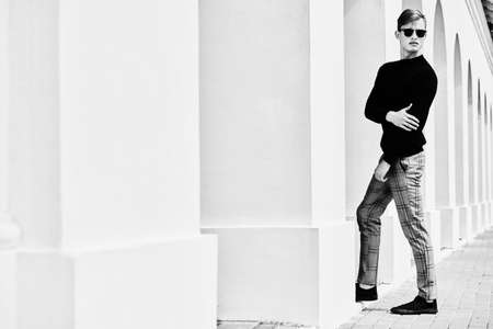 Fashion shot. Black-and-white portrait of a handsome young man model in black pullover, plaid trousers and black sunglasses posing next to white columns in the old town.の写真素材