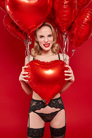 Love and Valentine's Day. Stunning blonde woman in sexual black lingerie poses with red heart shaped balloons on a red background.の写真素材