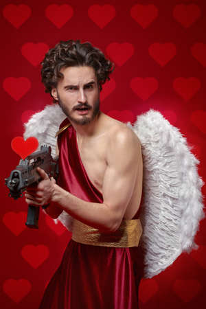 Modern Cupid with a big gun looking for a sacrifice of love. Red background. God of Love. Valentine's Day and Love.の写真素材