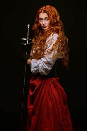 Beautiful courageous lady with long foxy hair posing with a battle epee (rapier) on a black background with fire sparks. Historical reconstruction of the 16-17th centuries. Copy space.の写真素材