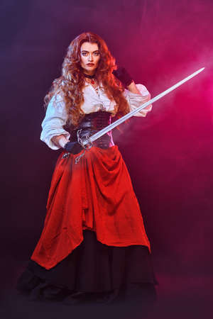 Beautiful redhead woman poses with a battle epee (rapier) on a black background with fuchsia light in the haze. Historical reconstruction of the 16-17th centuries.の写真素材