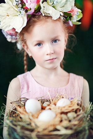 Portrait of redhead little girl with bunny ears holding nest with light eggs. Easter holiday....