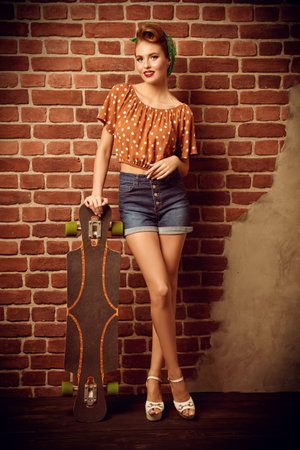 Attractive pin-up girl posing by the brick wall with her skateboard. Retro shooting.