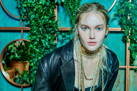 Stylish young woman model poses in greenhouse in black leather jacket and gold and pearl jewelry....