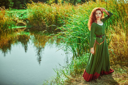 Portrait of a beautiful young woman archer with magnificent long red hair in a historical celtic dress is posing near the lake. Historical reconstruction.の写真素材