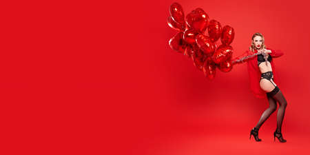 Stunning blonde woman in sexual black lingerie posing with red heart shaped balloons on a red background. Copy space. Love and Valentine's Day concept.の写真素材