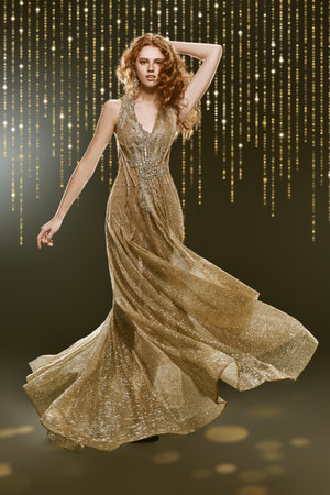 Full length portrait of a beautiful red-haired woman in a luxury golden dress in motion surrounded by festive golden lights. Beauty, evening fashion.の写真素材