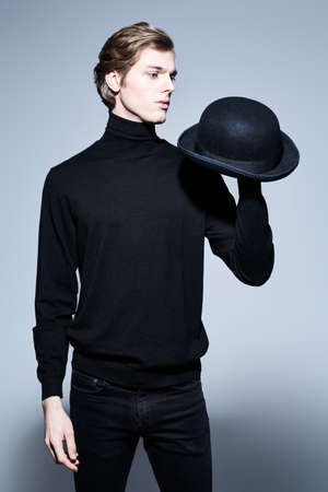 Portrait of an elegant young man posing in a black turtleneck and with a bowler hat on a gray background. Men's beauty. Fashion.の写真素材