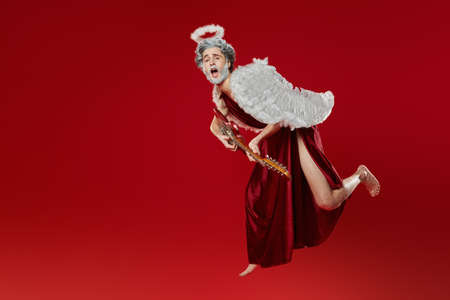 Portrait of emotionally singing Cupid flying with an electric guitar. Contemporary rock musician Amur. God of Love. Valentine's Day and Love. Studio portrait on a red background.の写真素材