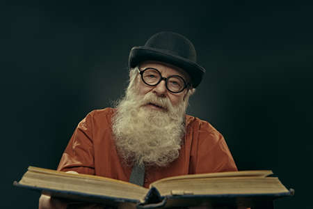 An intelligent old man in glasses and bowler with a long gray beard holds an old big book and looks at the camera. Old age wisdom.の写真素材