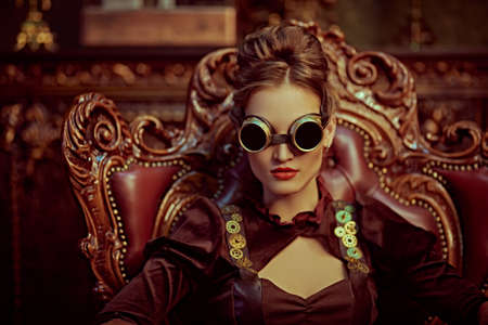 Beautiful steampunk woman with a glasses sits in an armchair in a room with vintage victorian interior. Science fiction, adventures.の写真素材