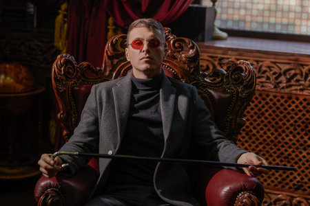 Imperious mature man in stylish expensive clothes and glasses sits in the armchair in the luxury...